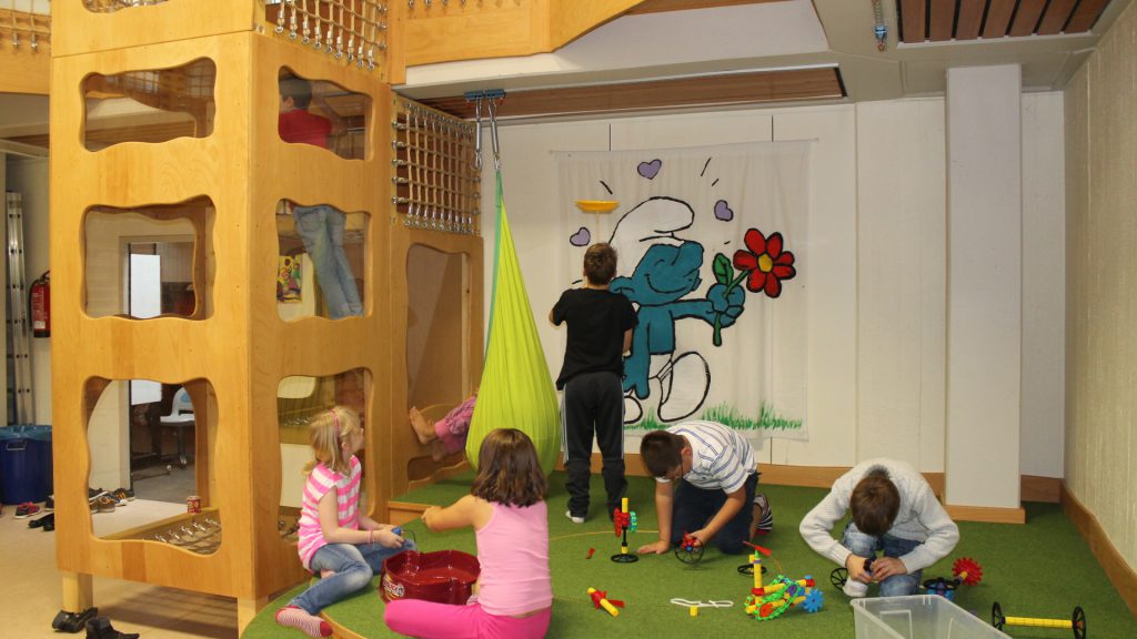 Sieben Kinder im Kinderraum des Jugendzentrums, mit Spielsachen auf einem Spielteppich oder den hölzernen Kletterturm hochkletternd.