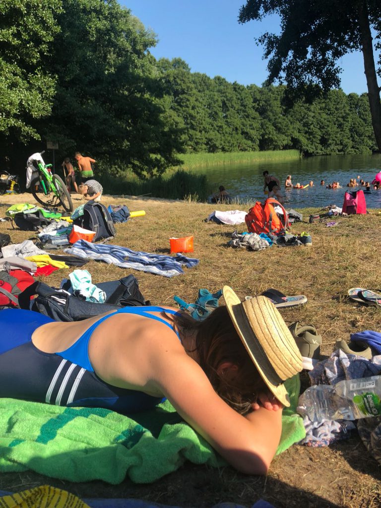 Ein Seeufer an dem diverse Handtücher und Rucksäcke liegen. Im Hintergrund sind menschen im Wasser.