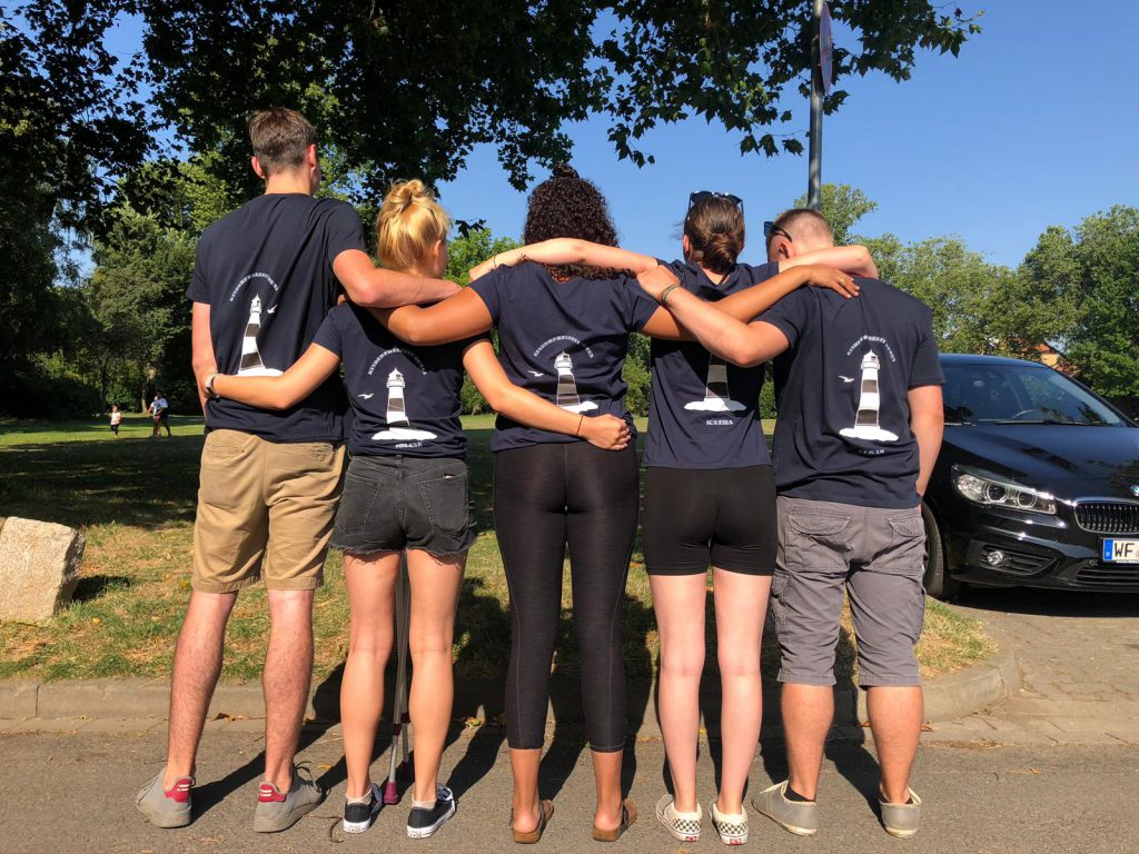 Fünf Jugendliche, die die Arme umeinander gelegt haben von hinten. Alle mit blauen T-shirts auf denen ein Leuchtturm abgebildet ist.