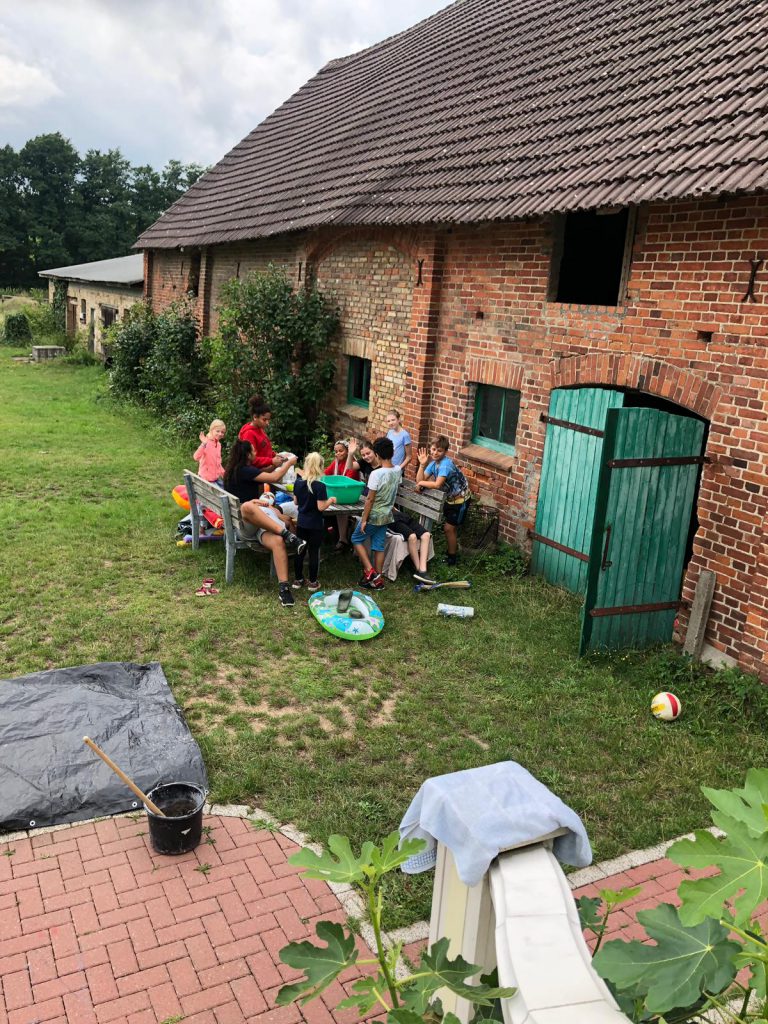 Mehrere Kinder auf Bänken an einem Tisch neben einem alten Bauernhaus.