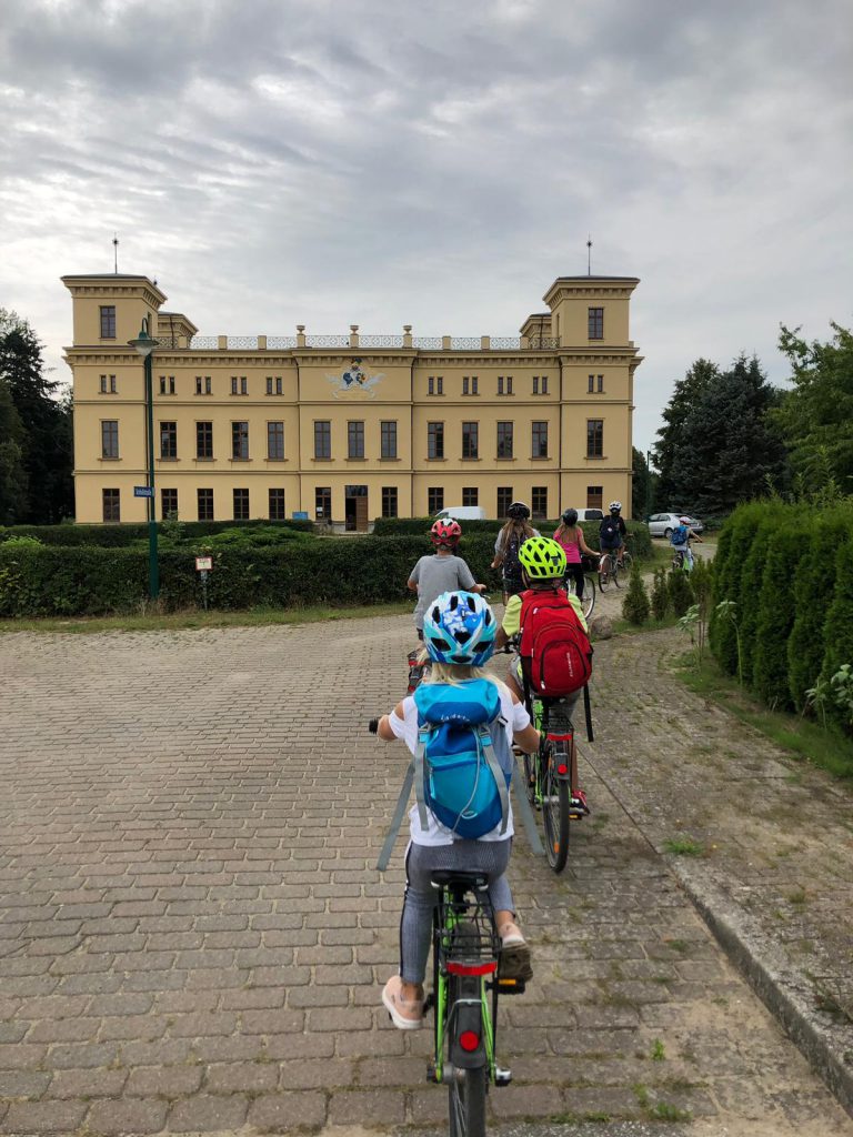 Mehrere Kinder auf Fahrrädern vor einem großen, alten Gebäude.