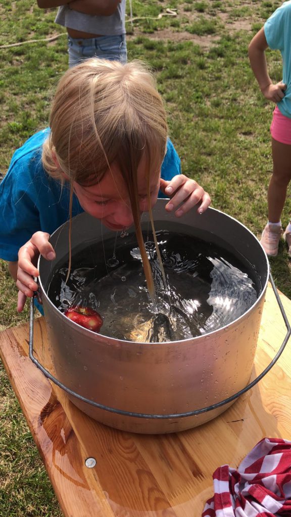 Ein Kind mit nassem Gesicht vor einem Topf mit Wasser, in dem ein Apfel schwimmt.