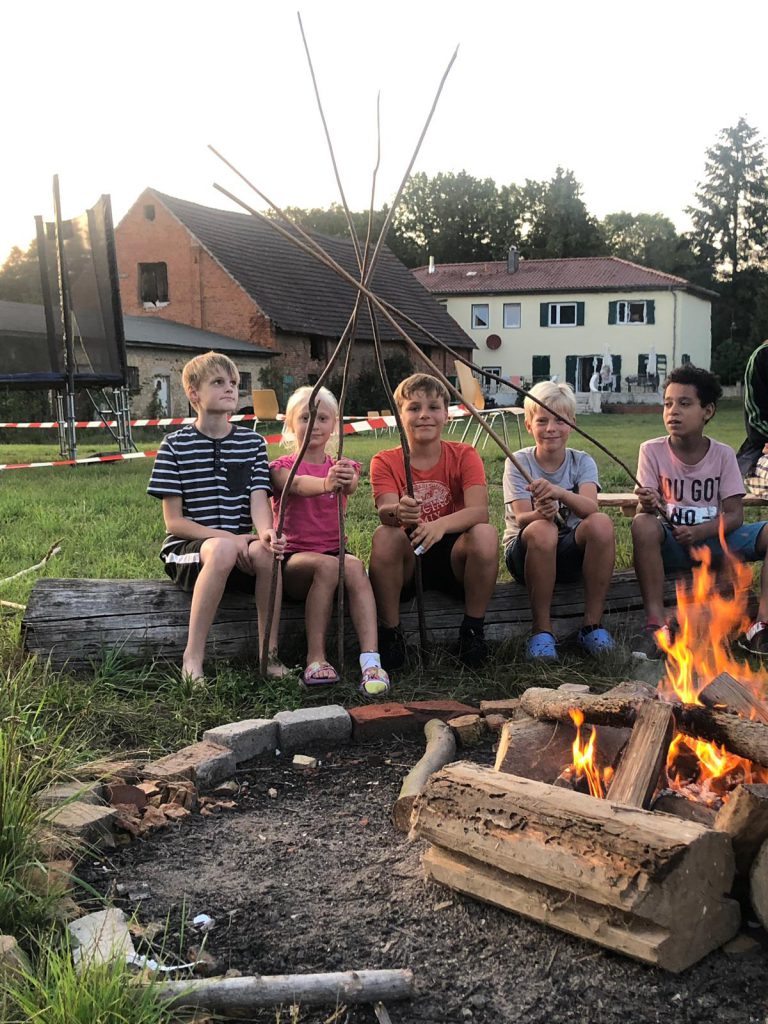 Fünf Kinder um ein Lagerfeuer mit langen Stöcken in der Hand.